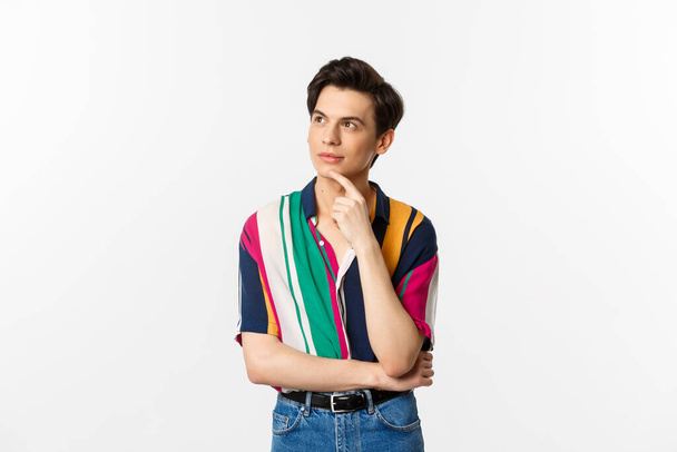 Portrait of thoughtful queer man in stylish shirt, looking at upper left corner and thinking, making choice, standing over white background - Foto, immagini