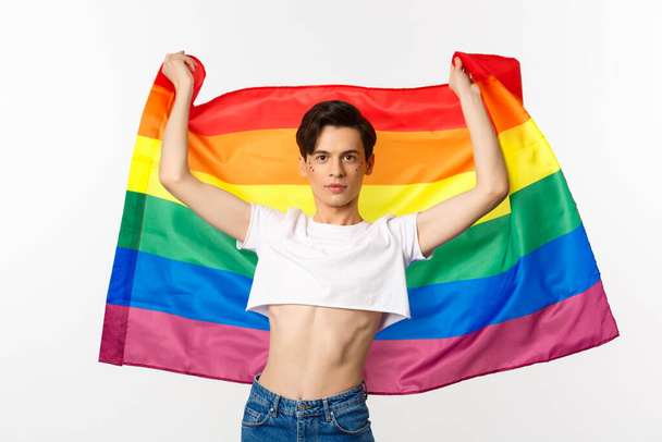 Młody człowiek w plonach, z brokatem na twarzy, wznoszący dumę z pewnością siebie. queer osoba uśmiecha się z flagą lgbt - Zdjęcie, obraz