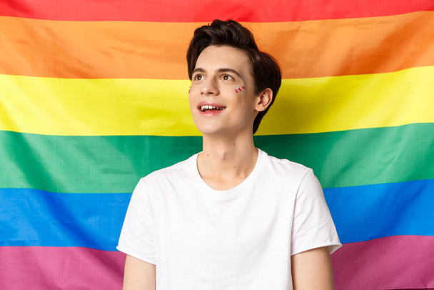 Waist-up shot of happy gay man with glitter on face smiling, looking dreamy at upper left corner, celebrating pride holiday, fighting for visibility, standing against lgbtq rainbow flag - Foto, imagen