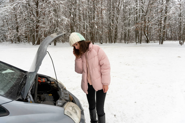 Jonge brunette vrouw in een roze winter donsjas en warme hoed staat in de buurt van een auto met een open motorkap en praten aan de telefoon - Foto, afbeelding