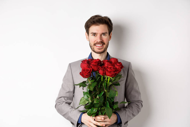Handsome young man in suit standing with gift on valentines day, holding red roses bouquet and smiling at lover, standing over white background - Fotó, kép