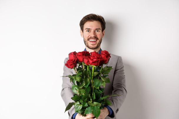 Schöner verliebter Mann, der einen glücklichen Valentinstag wünscht, beim romantischen Date einen Blumenstrauß schenkt, in die Kamera lächelt, Anzug über weißem Hintergrund trägt - Foto, Bild