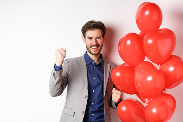 Lovers day. Cheerful young man in suit, feeling confident about Valentines date, saying yes and smiling, standing near hearts balloon, white background - Photo, Image