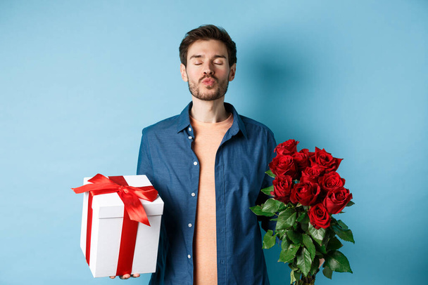 Láska a Valentýn koncept. Romantický muž čeká na polibek, drží dárkové krabice a kytice rudých růží pro milence na rande, stojící nad modrým pozadím - Fotografie, Obrázek