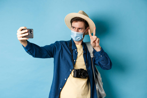 Covid-19, pandemic and travel concept. Счастливый и позитивный парень в туристической шляпе, делающий селфи и показывающий знак мира, позирующий рядом с достопримечательностями, на голубом фоне - Фото, изображение