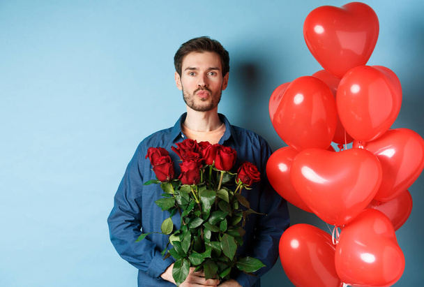 Romantic man with red roses and red heart balloons, pucker lips for kiss, making surprise on Valentines day, standing against blue background - Photo, image