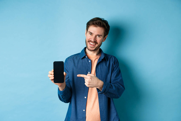Homme caucasien souriant montrant sur l'écran vide du smartphone, pointant vers le téléphone mobile et regardant satisfait, debout sur fond bleu - Photo, image
