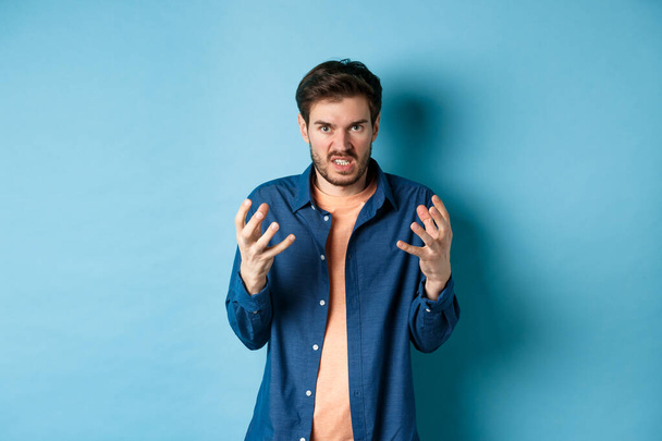 Annoyed and angry man raising hands and clench teeth outraged, stare pissed-off at camera, going to kill someone, standing on blue background - Foto, afbeelding