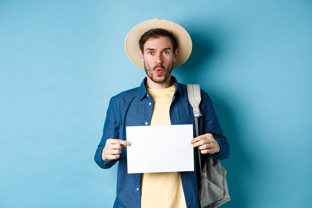 Aufgeregte Touristin mit Strohhut, leerem Blatt Papier in der Hand und amüsiert guckend, auf Sommerreise, auf blauem Hintergrund stehend - Foto, Bild