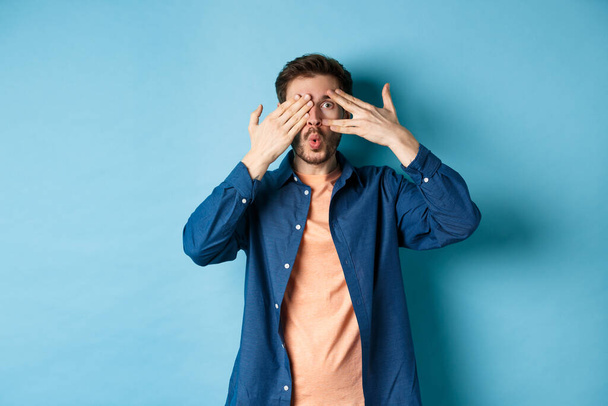Excited young man peeking through fingers and saying wow, checking out surprise gift, standing on blue background - Фото, изображение