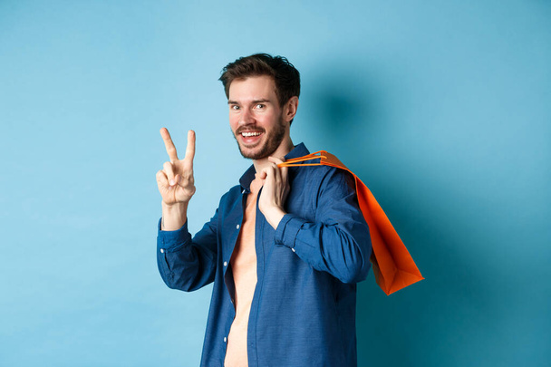 Gai gars tenant sac orange shopping sur l'épaule, souriant et montrant signe de paix, debout sur fond bleu - Photo, image