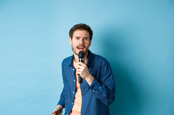 Страстный певец смотрит в камеру, поет в микрофоне, играет в караоке на синем фоне - Фото, изображение
