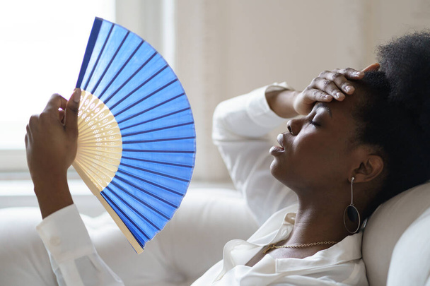 Afro γυναίκα των επιχειρήσεων που πάσχουν από θερμοπληξία κάθεται στο σαλόνι στο σπίτι χρησιμοποιώντας κυματίζει ανεμιστήρα - Φωτογραφία, εικόνα