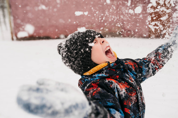 niño preescolar lanza nieve y está muy feliz por el invierno en ropa de abrigo, sombrero, snood amarillo, retrato infantil de la vida real de cerca, enfoque selectivo - Foto, Imagen