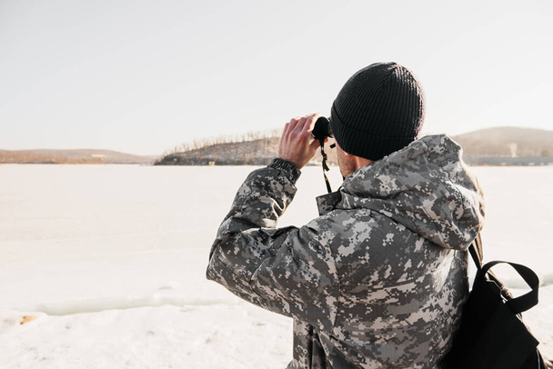 hunter with bristles in military clothes and a blue hat looks through binoculars, winter wildlife solo traveling, selective focus - Photo, Image