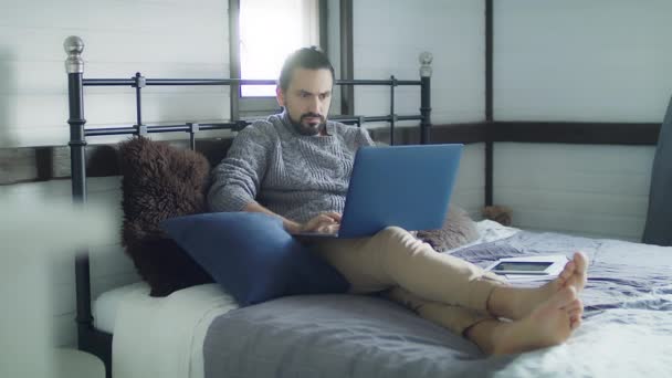 ベッドで寝そべっている間にノートパソコンで働く若いハンサムな男。ソーシャルメディアには居心地の良いグレーのニットセーターの男が立ち往生しています。若い男がネットサーフィンをしてる. - 映像、動画