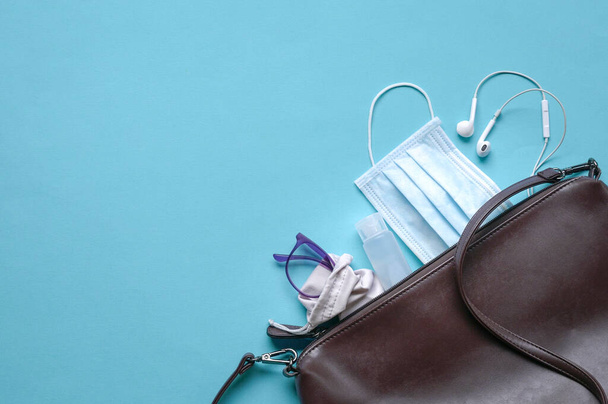Woman pouch with face mask, sanitizer, eyewear and earphones on blue background. Health and safety on the move concept. - Valokuva, kuva