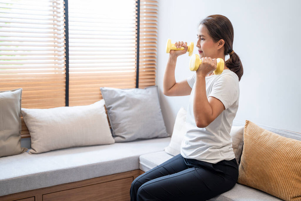 Mujer asiática joven atractiva y saludable haciendo ejercicio en casa durante el entrenamiento con ropa cómoda en una alfombra en su sala de estar, concepto de estilo de vida saludable. - Foto, Imagen