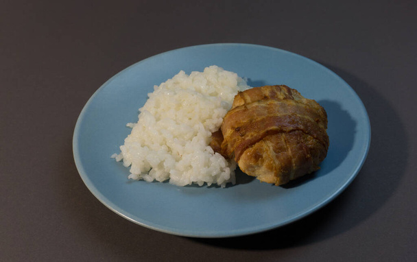 Főtt fehér rizs csirke szelettel, szalonnába csomagolva, szürke háttér kék tányéron. Sült húsgombóc körettel. - Fotó, kép