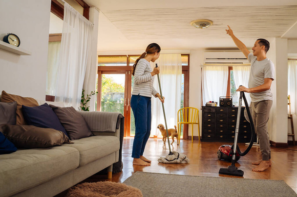 Happy young couple of woman dancing and singing while cleaning with vacuum cleaner machine and man mopping the floor in living room at home - Φωτογραφία, εικόνα