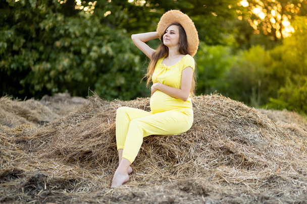 een zwangere vrouw in donkere kleren en een hoed zit in een veld op stro in de zomer - Foto, afbeelding