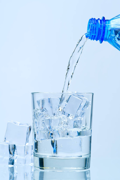 Выливание чистой питьевой воды из голубой пластиковой бутылки в стекло на голубом фоне - Фото, изображение