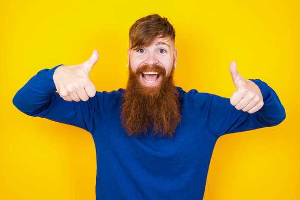 A boldog mosolygós, jóképű, vörös hajú, szakállas fehér férfi portréja, aki a sárga falnál áll, és két hüvelykujjával felfelé mutat: Jó munka! Pozitív emberi érzelmek. - Fotó, kép