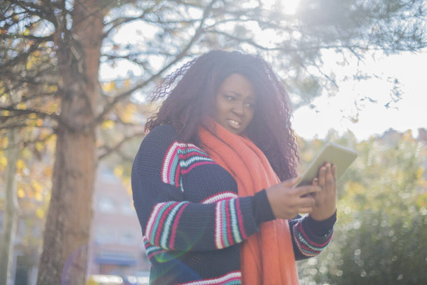 beautiful african american woman wearing sweater and orange scarf holding  tablet computer   in autumn  park   - Photo, Image