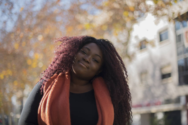 若い美しいアフリカ系アメリカ人女性は,秋の公園でカメラを見ているセーターとスカーフを着ています    - 写真・画像