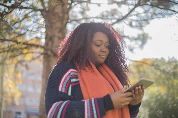 young beautiful african american woman wearing sweater and scarf  using tablet  in autumn  park    - Foto, Imagen