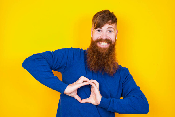 красивий рудий бородатий кавказький чоловік стоїть на тлі жовтої стіни, посміхаючись у коханні, демонструючи серцевий символ і форму руками. Романтична концепція. - Фото, зображення