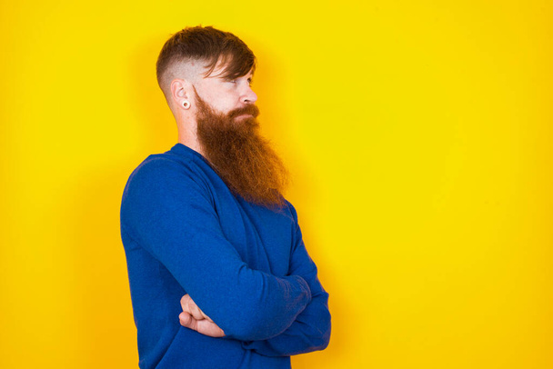 Image of upset handsome red haired bearded Caucasian man standing against yellow wall  with arms crossed. Looking with disappointed expression aside after listening to bad news. - Photo, Image
