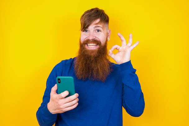 Счастливый красивый рыжеволосый бородатый белый мужчина стоит напротив желтой стены, посылая сообщение на свой смартфон или делая селфи и делая хорошо знак рукой. - Фото, изображение