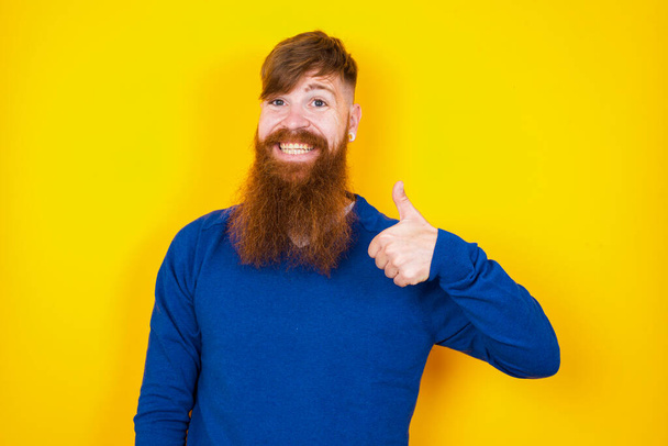 schöner rothaariger Mann mit kaukasischem Bart, der an der gelben Wand steht und Daumenhoch-Geste macht, guter Job! Positive menschliche Emotion Gesichtsausdruck Körpersprache. - Foto, Bild