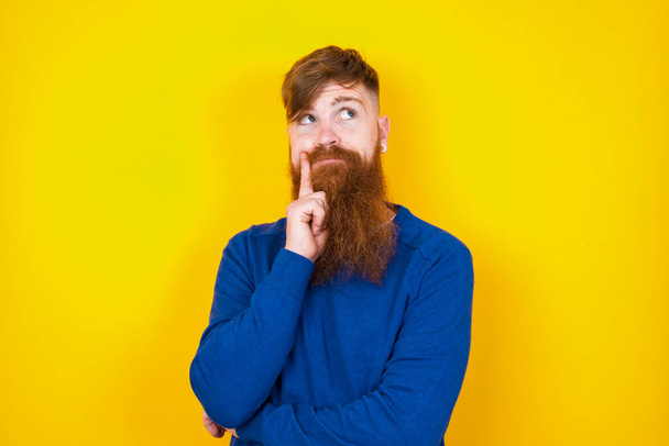 Portrait of thoughtful handsome red haired bearded Caucasian man standing against yellow wall keeps hand under chin, looks away trying to remember something or listens something with interest. Youth concept. - Photo, Image