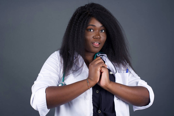 Dreamy young female doctor with pleasant expression keeps hands crossed near face, excited about something pleasant, poses against gray background. Young African woman wearing medical white gown - Foto, Bild