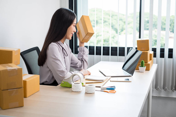 Young Asian woman business owner with many parcel boxes on the table happy online sales job, use your laptop, get an order from customers, take notes, and make arrangements for delivery by post. - Photo, Image