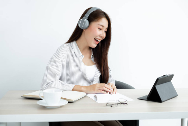 Video Llamar a una joven asiática con auriculares se comunica a través de vídeo en línea de larga distancia en una tableta digital felicitaciones felices con un amigo. - Foto, Imagen