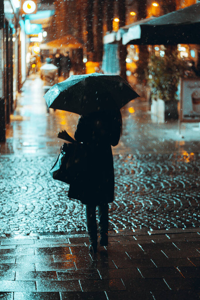 A back view of a woman wearing a black coat walking on the street holding an umbrella under the rain - Foto, Imagem