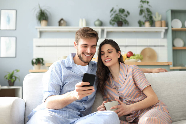 Young couple in pajamas watching online content in a smart phone sitting on a sofa at home in the living room - Fotografie, Obrázek
