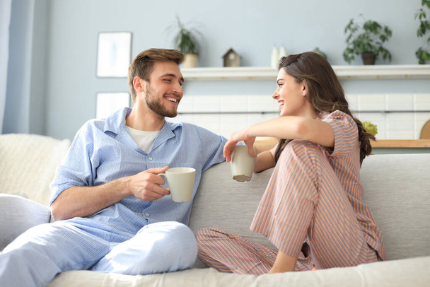 Beautiful young couple in pajamas is looking at each other and smiling on a sofa in the living room - 写真・画像