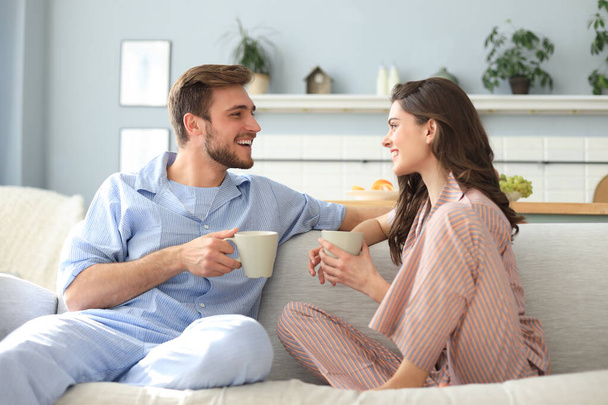 Beautiful young couple in pajamas is looking at each other and smiling on a sofa in the living room - Foto, imagen