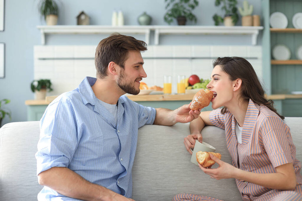 Happy young couple in pajamas in kitchen having breakfast, feeding each other a croissant - Фото, изображение
