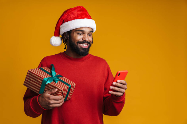 Happy african american guy using cellphone and holding Christmas gift isolated over yellow background - Foto, afbeelding