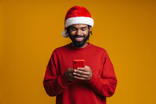 Happy african american guy in Santa Claus hat using cellphone isolated over yellow background - Foto, Imagem