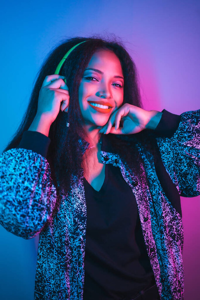 A portrait of a smiling Hispanic attractive woman illuminated by neon lights listening to music on headphones - Foto, imagen