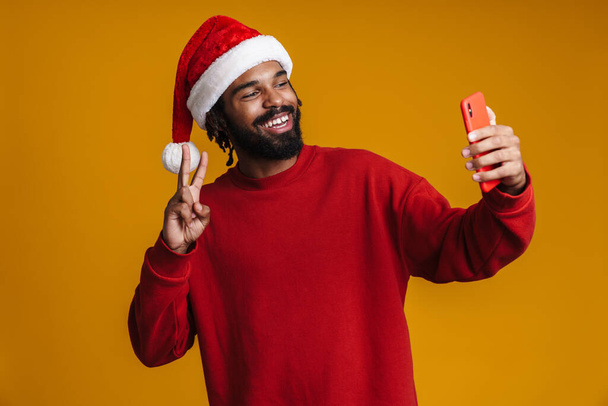 heureux afro-américain guy gestuelle signe de paix tout en prenant selfie sur téléphone portable isolé sur mur jaune - Photo, image