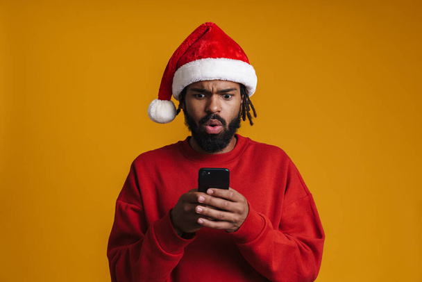 Brooding african american guy in Santa Claus hat using cellphone isolated over yellow background - Foto, Imagem