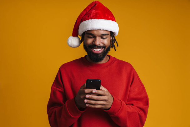 Happy african american guy in Santa Claus hat using cellphone isolated over yellow background - Φωτογραφία, εικόνα
