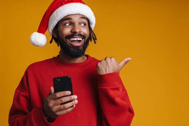 Happy african american guy pointing finger aside while using cellphone isolated over yellow background - Φωτογραφία, εικόνα